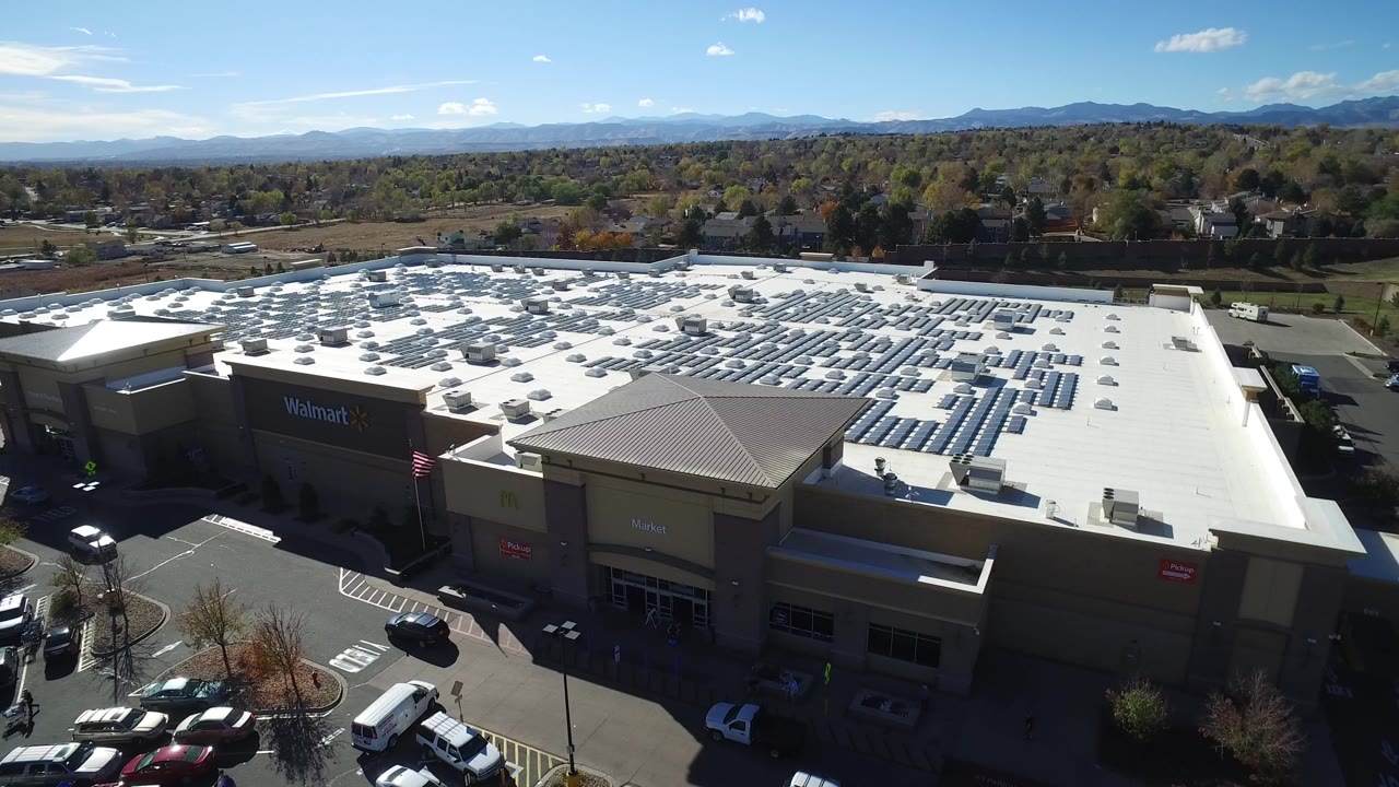 Aerial frame over Hidden Lake, Colorado showing a Walmart shopping mall roof with solar panels, parking areas, distant homes, mountains, and no visible people.
