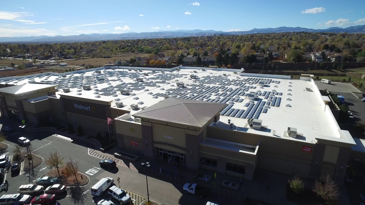 Elevated Hidden Lake, Colorado view with the mall roof centered, rows of solar panels across the white surface, cars near the storefront, and no visible people.