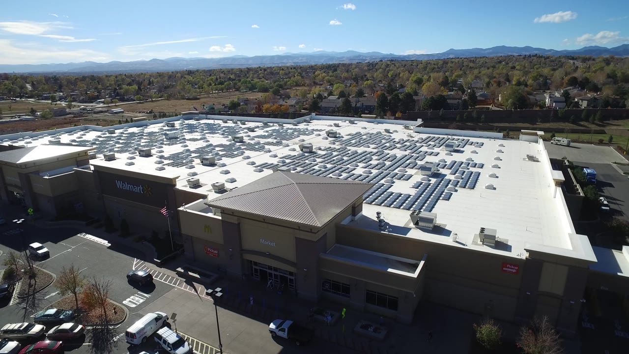 Aerial mall rooftop with solar panels, parked cars, trees, and distant mountains.