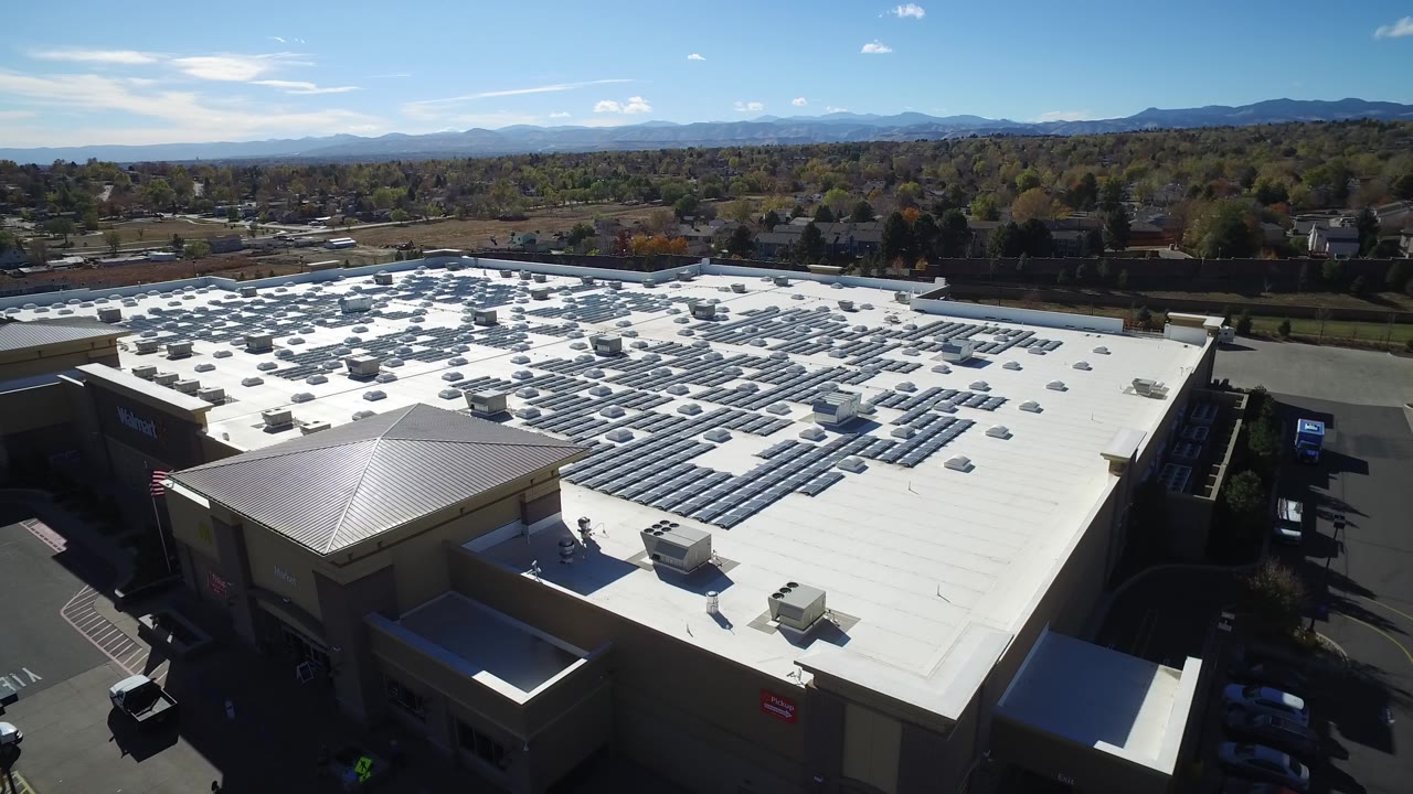 Aerial view over a large white-roofed commercial building with parking lots nearby.