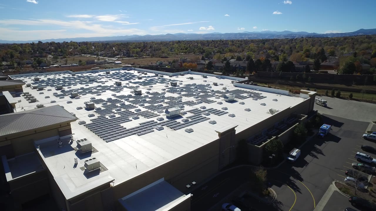 Elevated drone view over a white-roofed warehouse in Hidden Lake, Colorado, with rooftop solar panels, parking areas, trees, mountains on the horizon, and no visible people.
