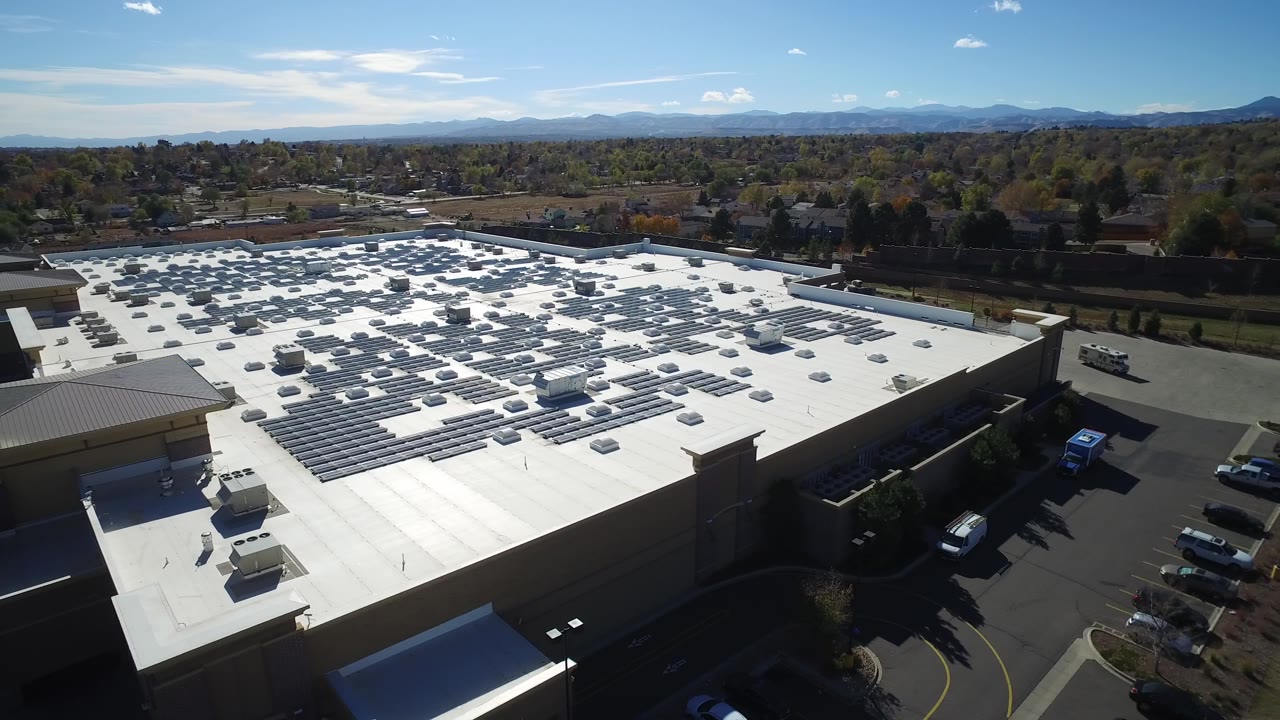 High angle view of a warehouse complex with solar panels, parking, trees, and mountains.