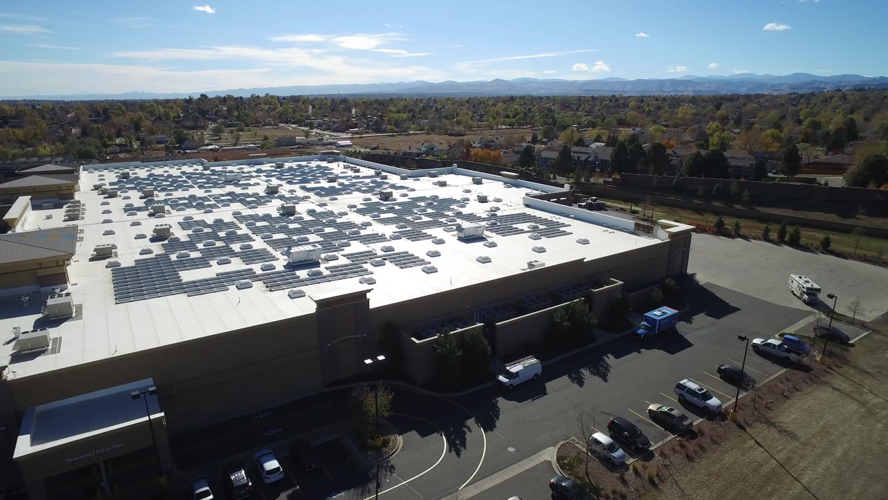 Aerial view of a large building roof, parking lot, cars, trees, and distant hills.