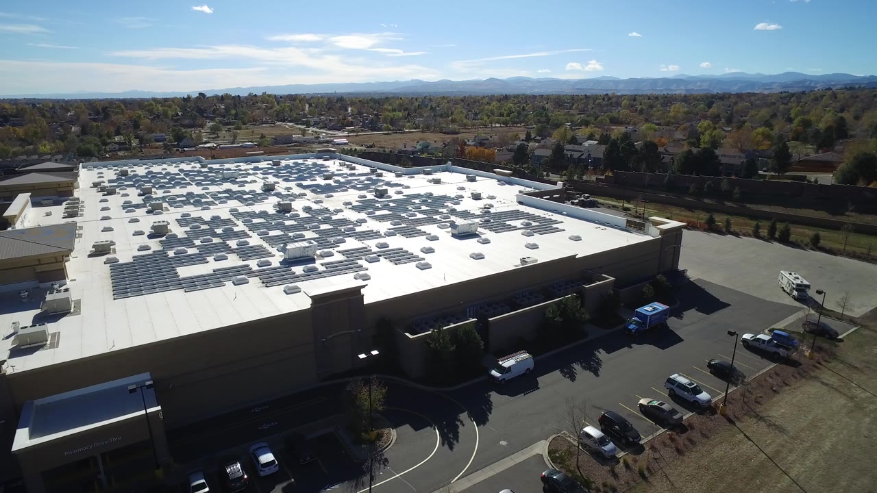 Aerial view in Hidden Lake, Colorado, centered on the white roof and parking rows, with suburban trees and low mountains in the background and no visible people.
