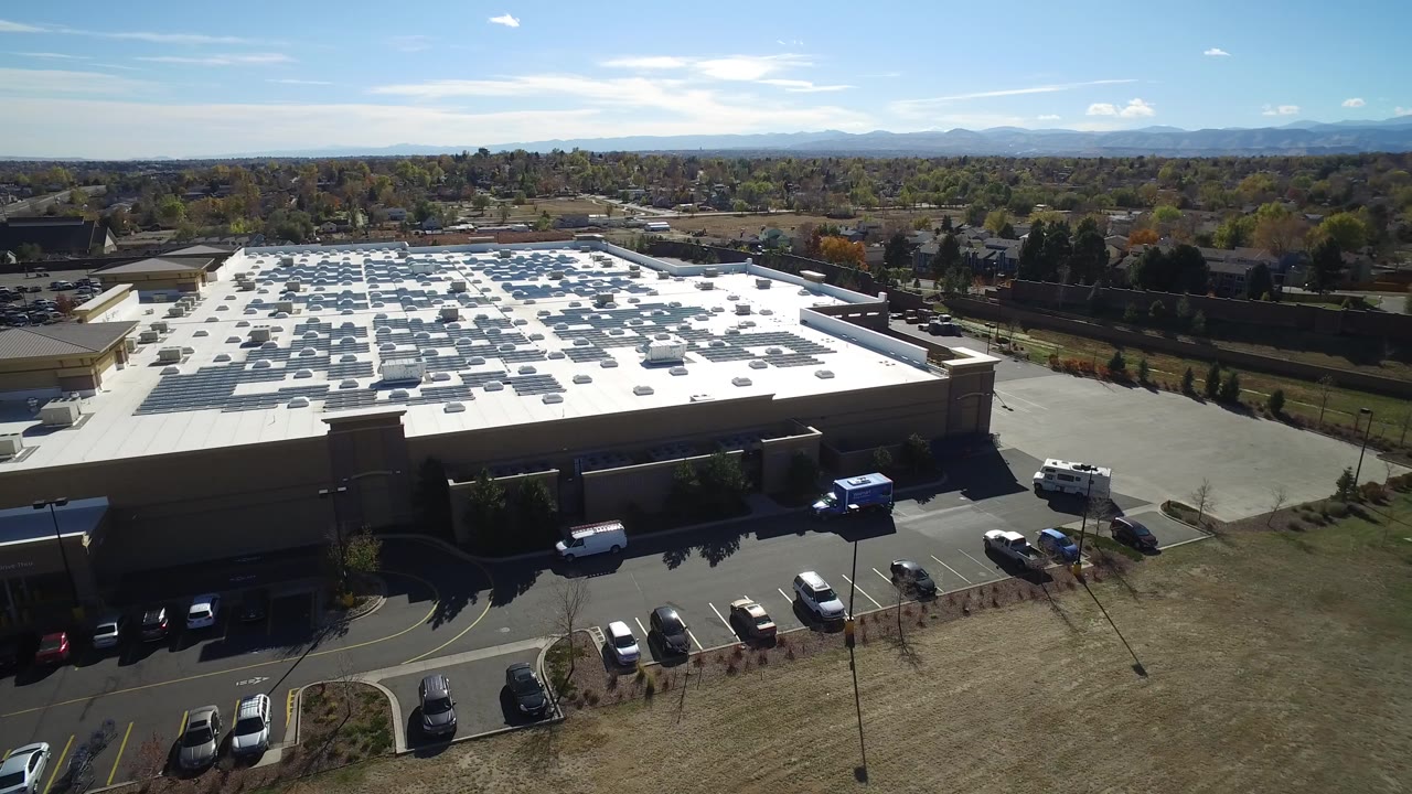 Wide aerial frame over Hidden Lake, Colorado showing a large tan commercial building, white roof equipment, parked cars, dry grass, distant neighborhoods, and no visible people.