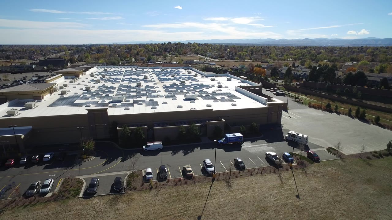 Aerial frame over Hidden Lake, Colorado showing a large flat-roofed commercial building, parked cars along the foreground lot, distant trees and mountains, and no visible people.