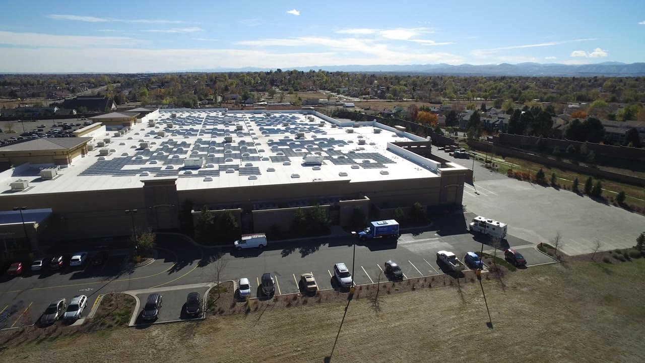 High aerial view in Hidden Lake, Colorado with the roof centered, rows of rooftop equipment visible, cars at the lower edge, low buildings beyond, and no visible people.