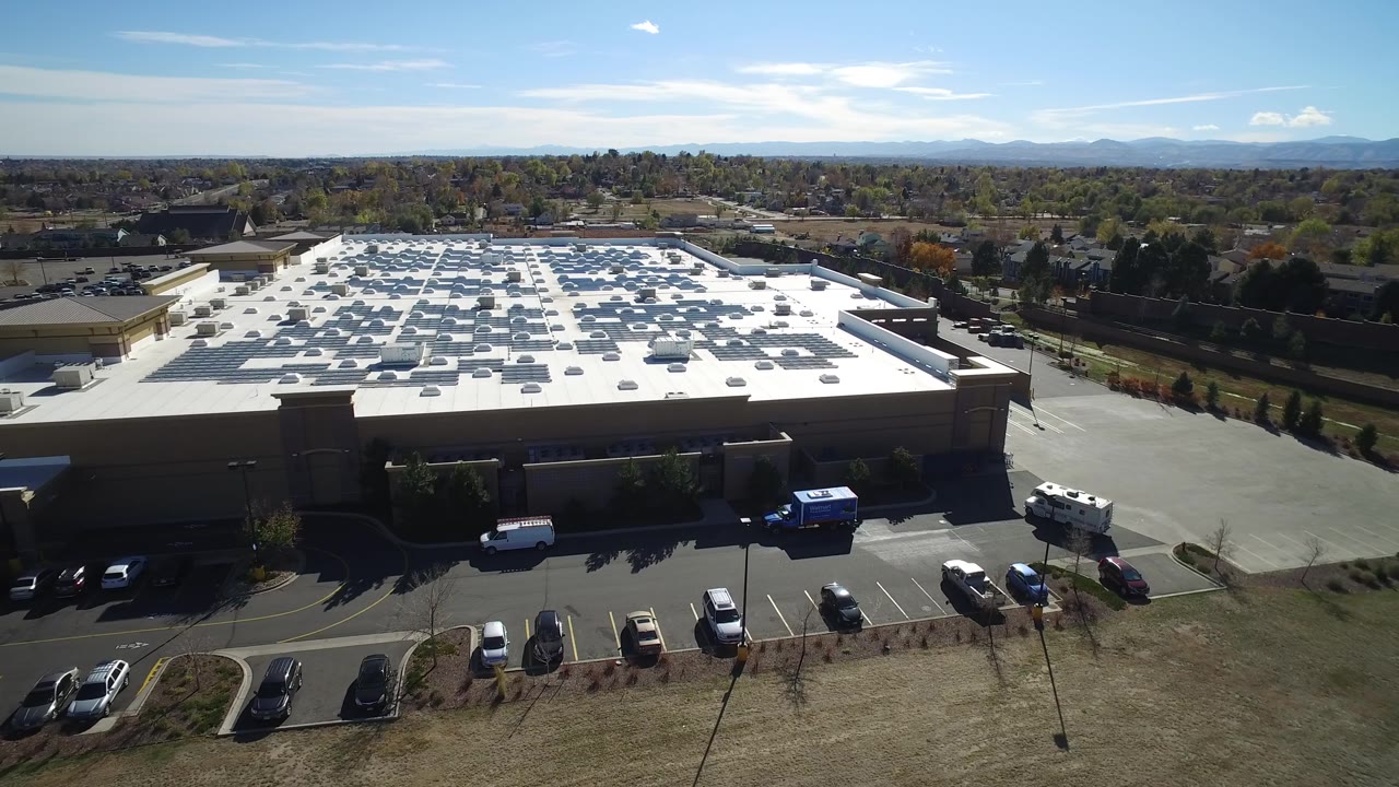 Drone-like view of a parking lot, building roof, and distant suburbs.