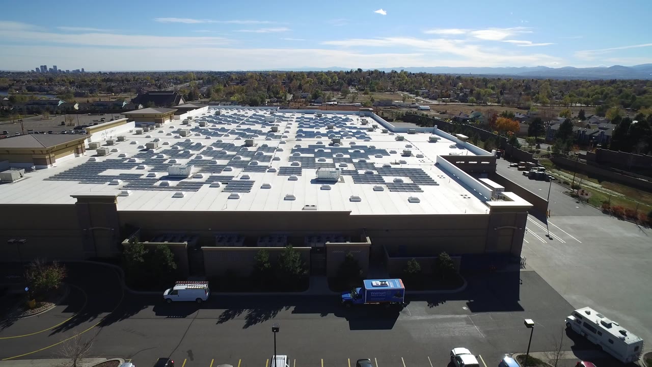 Elevated view over a large white-roofed commercial building in Hidden Lake, Colorado, with rooftop panels or units, service vehicles, trees, distant suburbs, and no visible people.