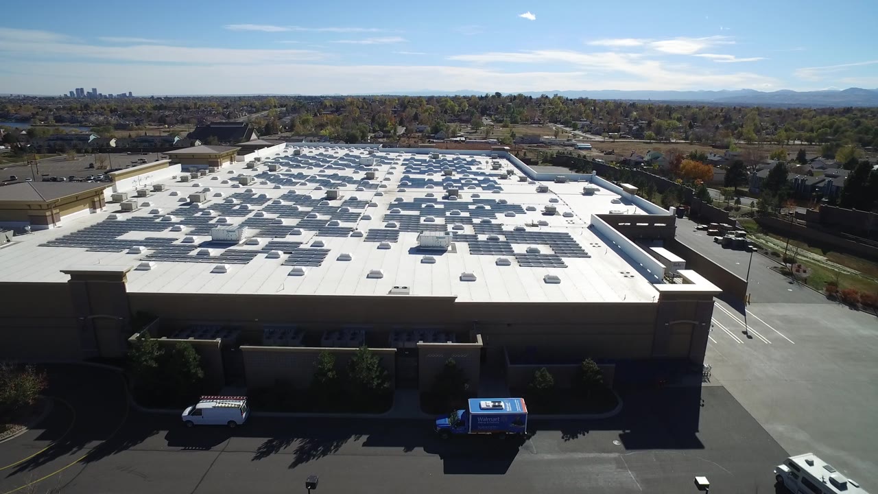 Aerial frame of a commercial roof with rooftop arrays and distant tree-lined suburbs.