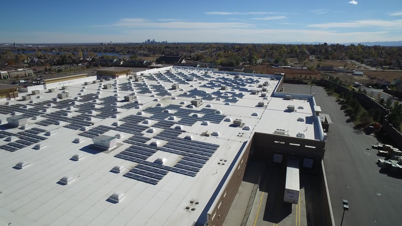Drone-height frame in Hidden Lake, Colorado with solar-panel rows across the roof, a paved lot on the right, distant houses and trees on the horizon, and no visible people.