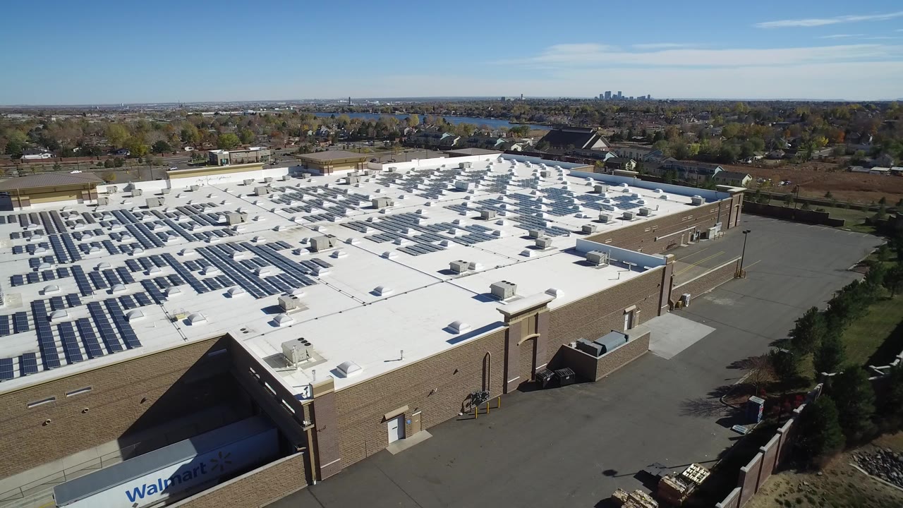 Aerial view in Hidden Lake, Colorado showing a white commercial roof lined with solar panels, tan exterior walls, service pavement, trees, distant homes, and no visible people.