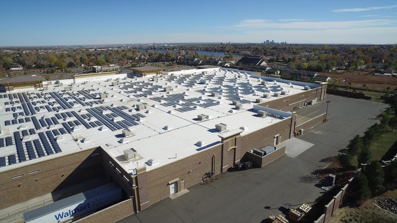 High aerial frame in Hidden Lake, Colorado with solar panels spread across the left side of the white roof, a tan building facade, asphalt service space, trees, distant residences, and no visible people.