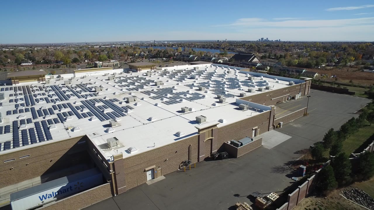 Drone-like view of a commercial rooftop solar array with homes and water in the distance.