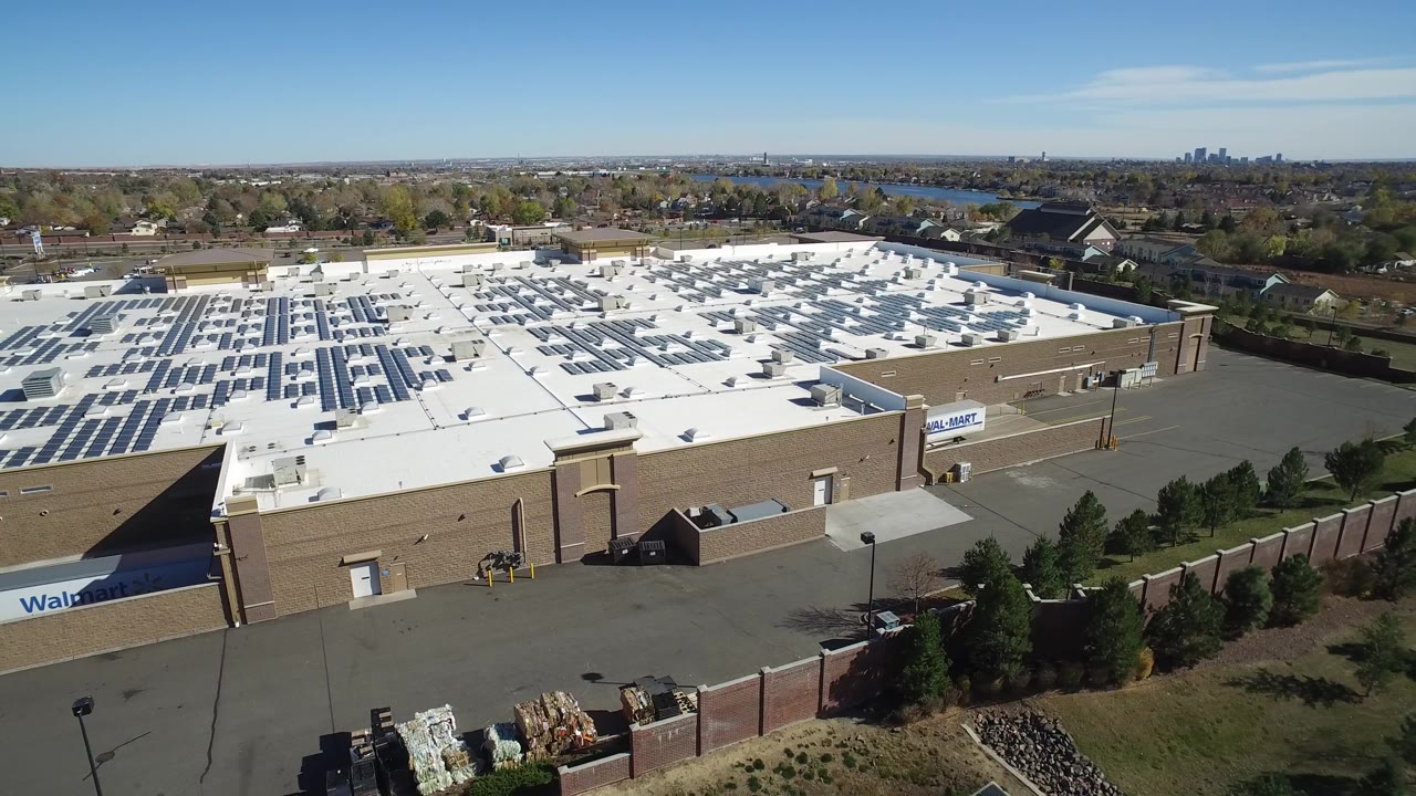 Aerial view in Hidden Lake, Colorado shows the retail building roof centered in frame, with solar-panel rows on the left side, rooftop vents, a service drive, trees, homes, water, and no visible people.