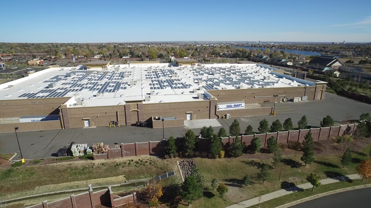 Aerial view of a large retail building with a white roof beside trees and a road.