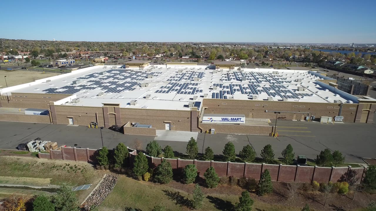 Elevated daylight view in Hidden Lake, Colorado of a large tan commercial building with a white roof, dark rooftop panels, paved service area, foreground wall and trees, and no visible people.