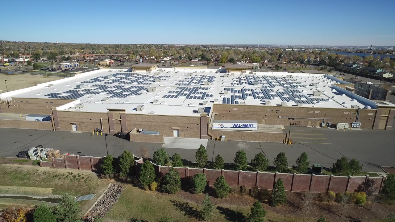 Drone-style daylight frame in Hidden Lake, Colorado showing the white-roofed commercial structure from slightly farther along the frontage, with rooftop equipment, background homes, and no visible people.
