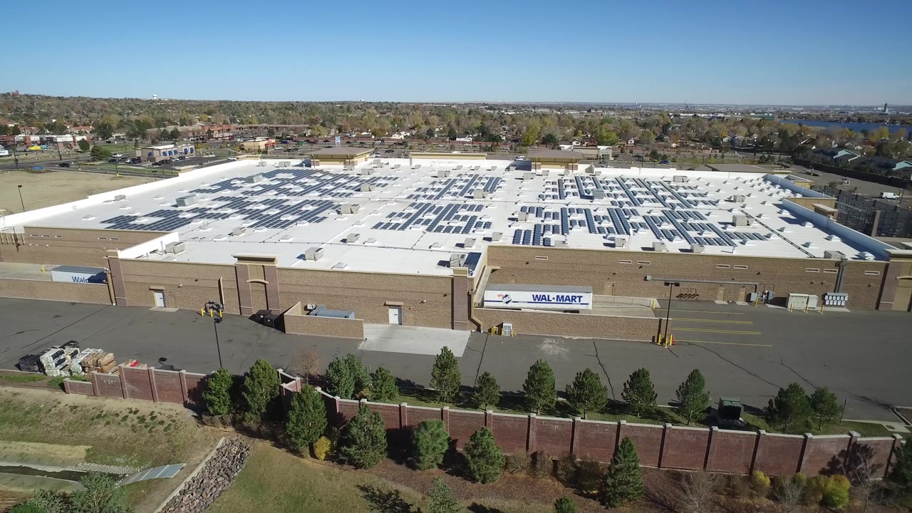 Wide aerial frame of a big-box commercial building with rooftop panels and a service road.