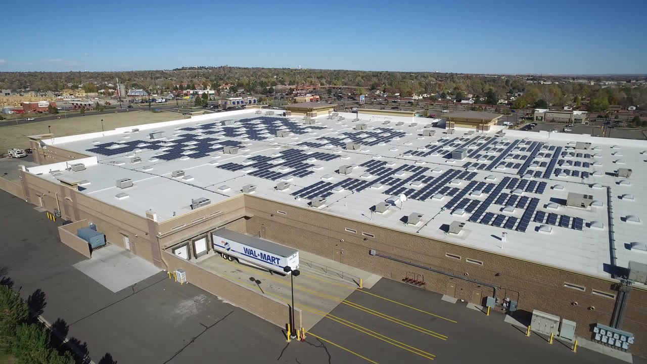 Second aerial frame in Hidden Lake, Colorado with the solar panel grid spread across the white roof, the loading dock edge and branded trailer lower in frame, and no visible people.