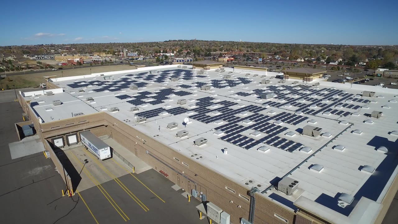 High aerial frame over a large shopping center in Hidden Lake, Colorado, showing white roofing, rows of dark blue solar panels, tan walls, loading docks, parking-lot markings, and no visible people.