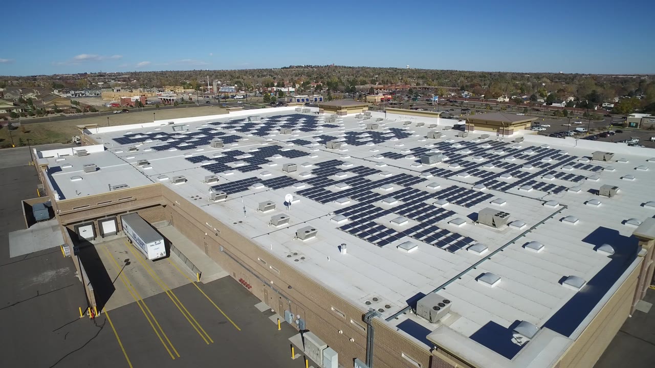 Wide aerial of a white-roofed shopping center with blue solar panels and nearby streets.