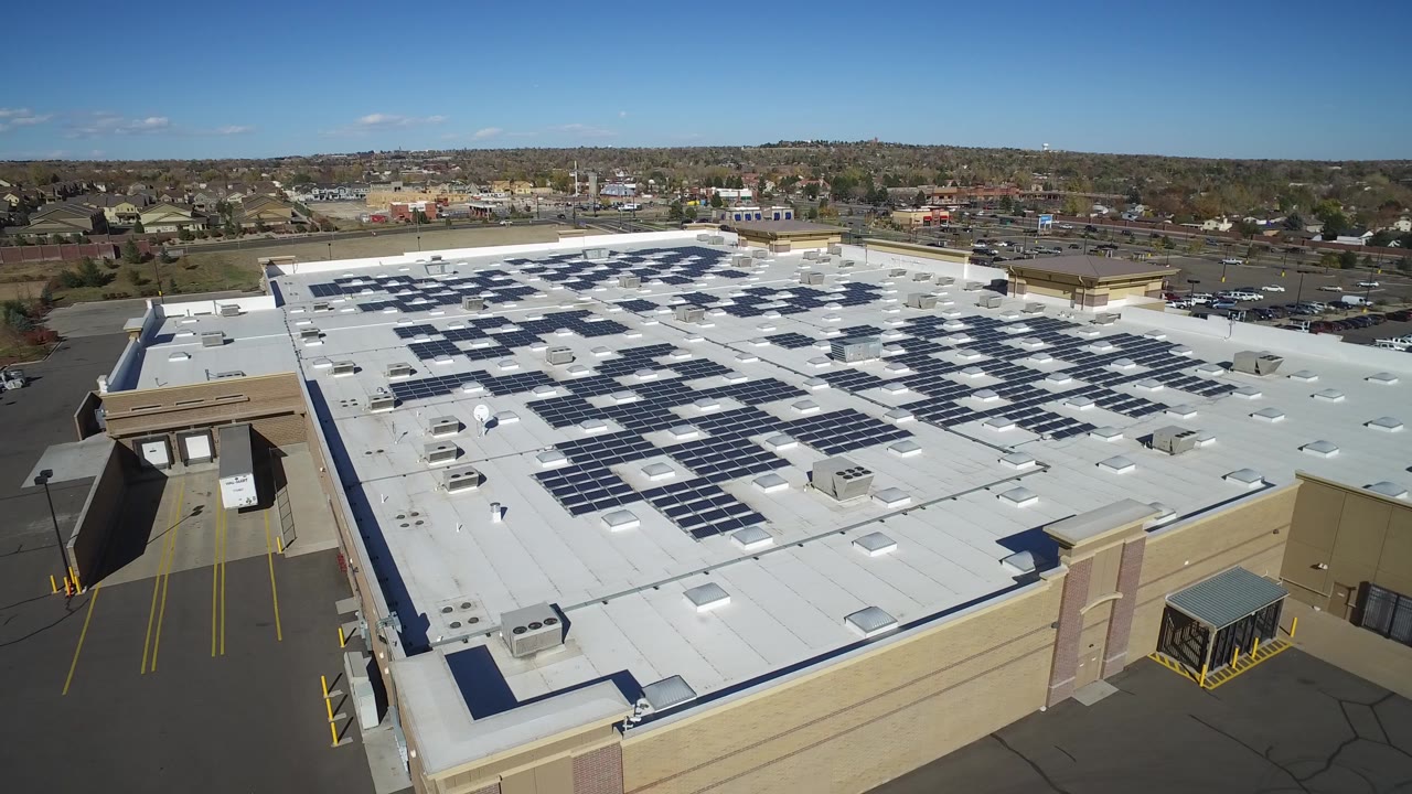 Aerial frame over Hidden Lake, Colorado showing a large white commercial roof with dark solar panel rows, rooftop vents, parking areas, distant buildings, and no visible people.