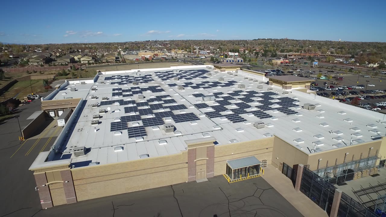 Opening aerial frame in Hidden Lake, Colorado showing a beige secure building with dark solar panels across a white roof, fenced pavement below, suburban development on the horizon, and no visible people.