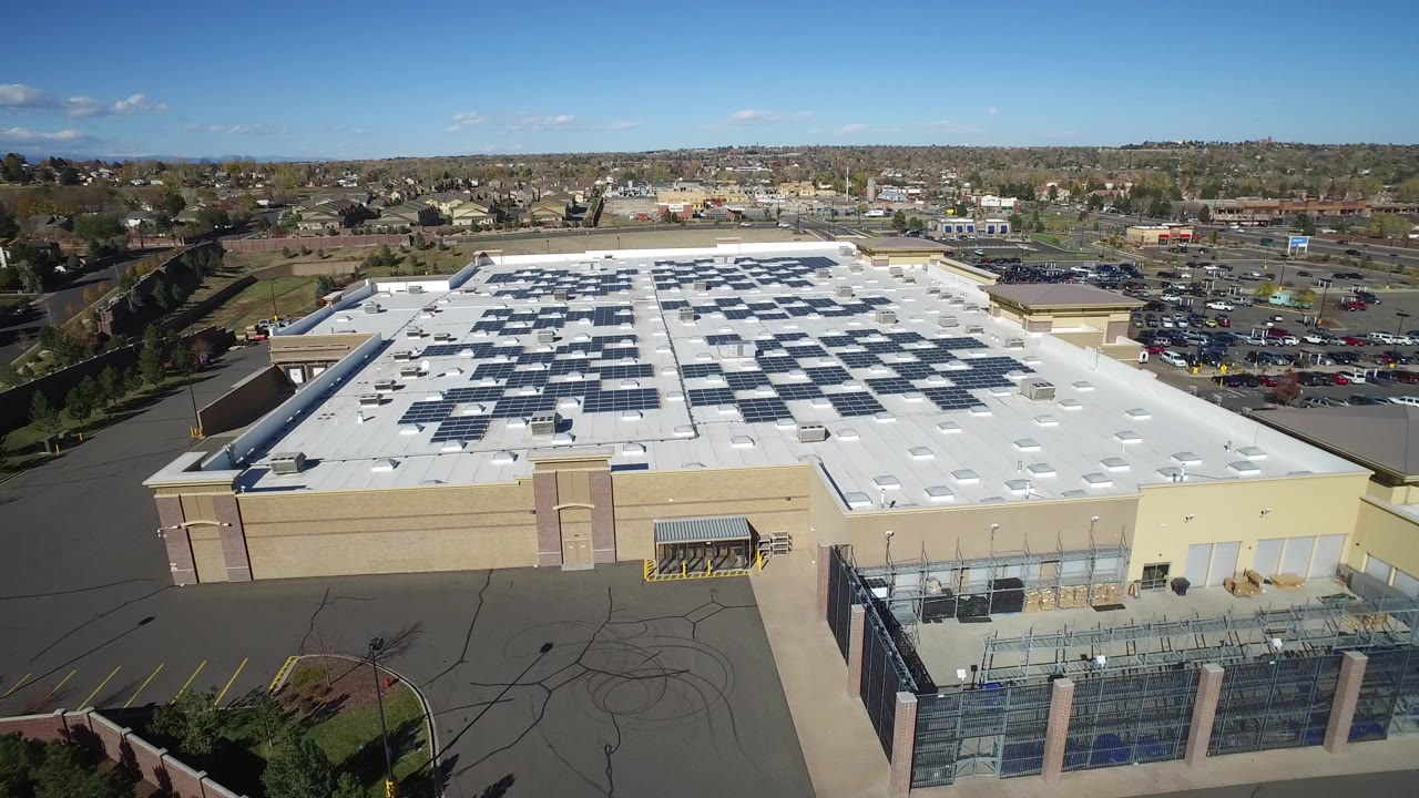 Opening aerial frame in Hidden Lake, Colorado showing a tan building complex with a white roof, solar panel arrays, fenced lower area, parking lots, trees, and no visible people.