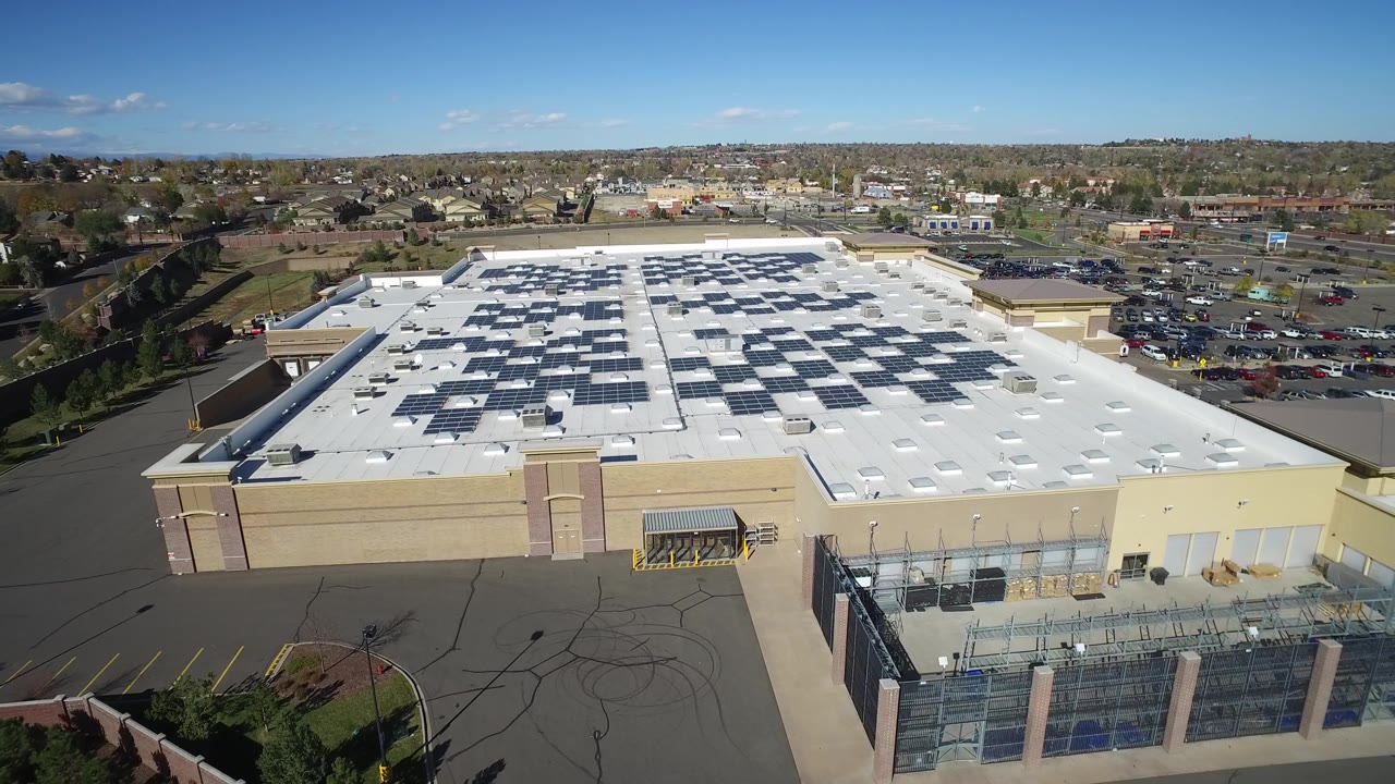 Second aerial frame in Hidden Lake, Colorado with the roof solar arrays centered, the building facade below, parked cars to the right, trees beyond, and no visible people.
