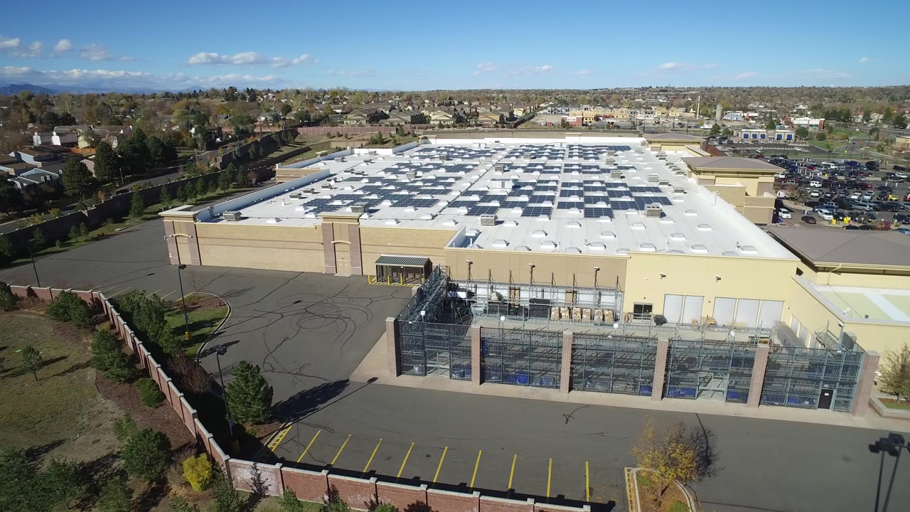 Aerial view of a beige commercial building, white roof, fenced structures, and parking lots.