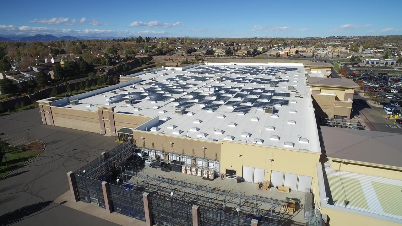 Aerial view of a large beige building with a white roof and fenced edge.