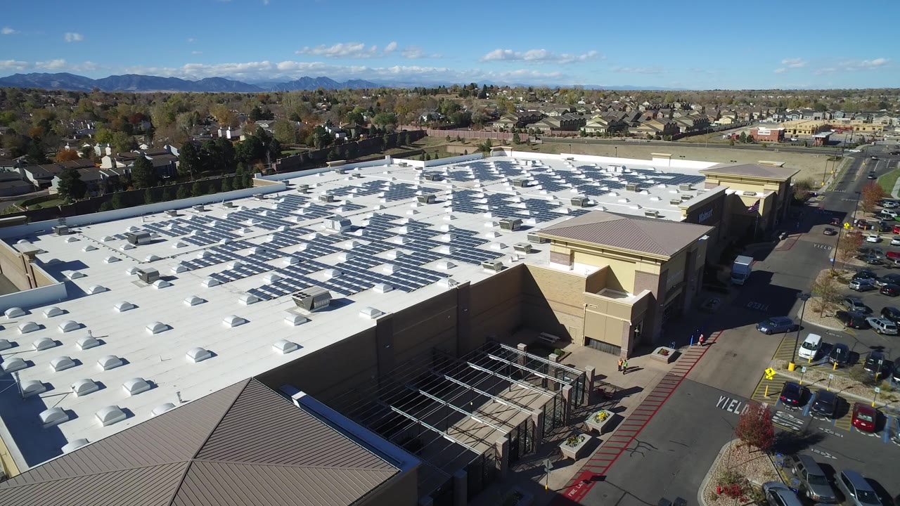 Aerial view over a mall roof, parking lot, homes, and distant mountains.
