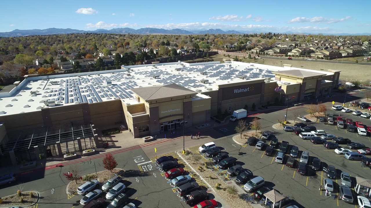Opening frame over Hidden Lake, Colorado shows a Walmart building with a white roof, parked cars, surrounding homes, distant mountains, blue sky, and no visible people.