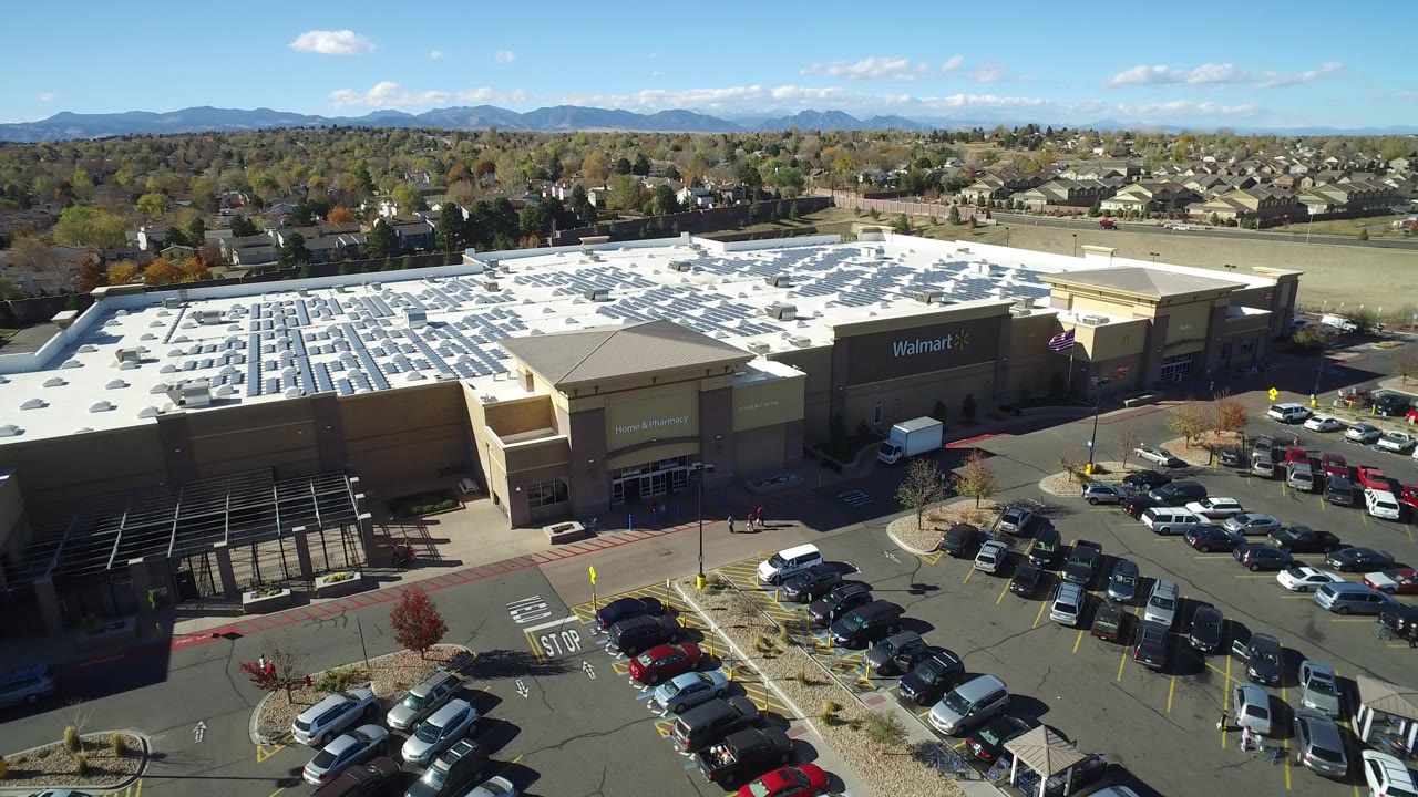 At 0:01, the Hidden Lake, Colorado aerial frame holds the large store roof in the center, with parking rows below, homes beyond the building, distant mountains, and no visible people.