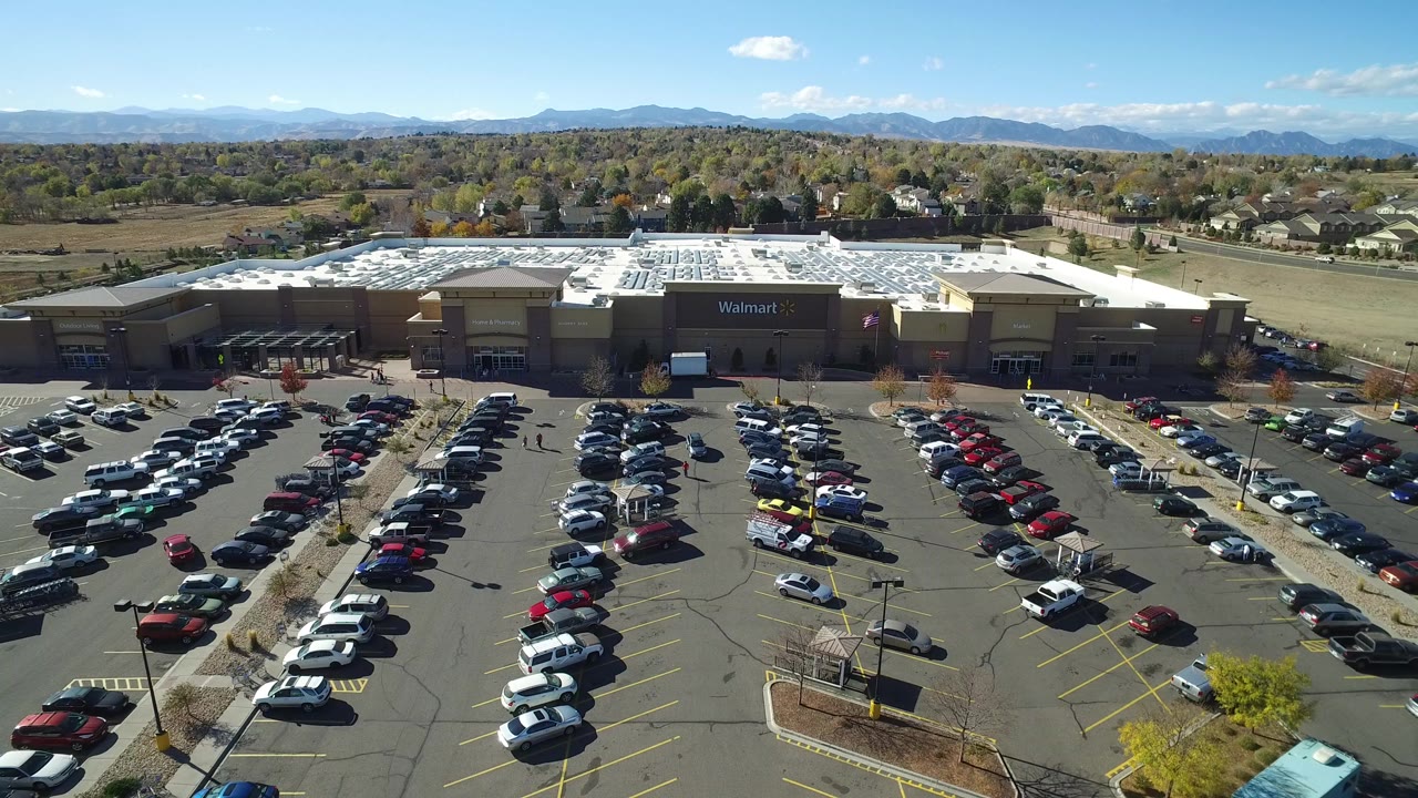 Opening aerial frame over a Hidden Lake, Colorado commercial parking lot, with parked cars in rows, a big-box storefront, distant homes and mountains, and no visible people.