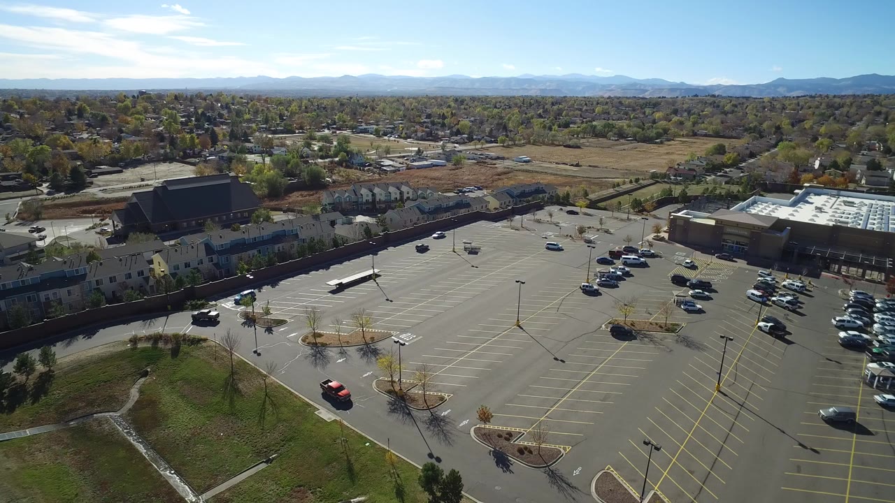 Aerial view over a large parking lot with cars, low buildings, trees, and distant mountains.