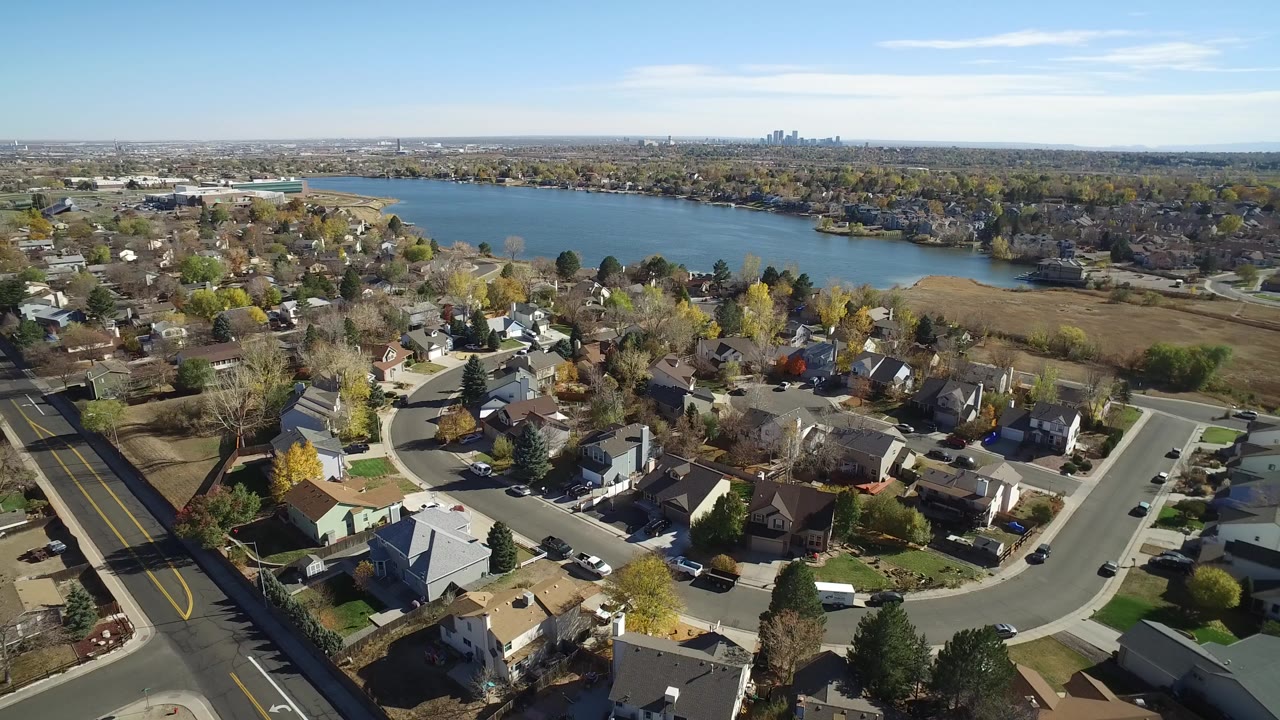 Opening aerial frame over Hidden Lake, Colorado, showing curved residential streets, detached homes, autumn trees, a blue lake, distant skyline, and no visible people.