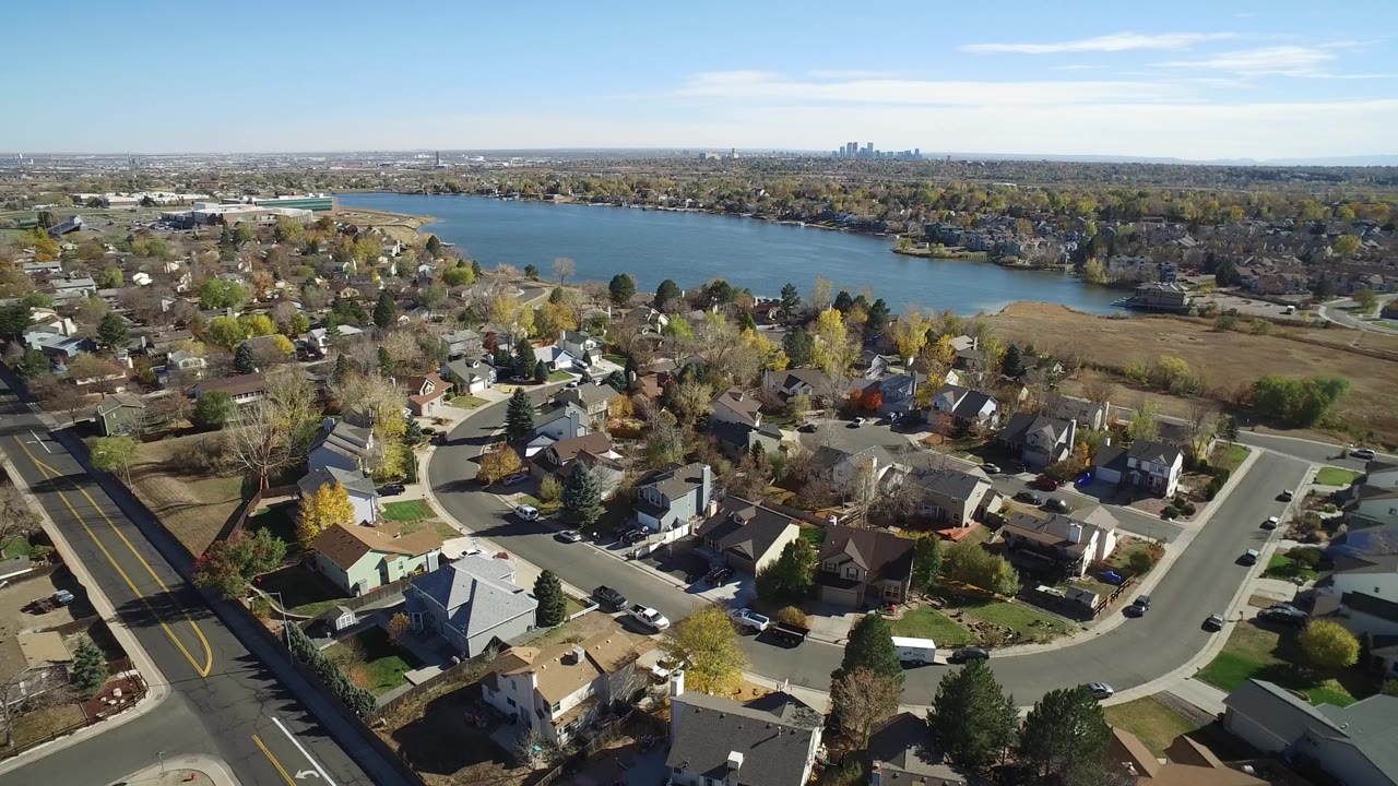 Aerial frame over Hidden Lake, Colorado, with a main road at left, clustered homes below, lake water across the upper frame, distant buildings on the horizon, and no visible people.