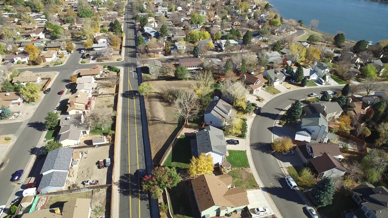 A high aerial view over Hidden Lake, Colorado shows a two-lane road beside detached homes and yards, with blue water at the top of frame and no visible people.