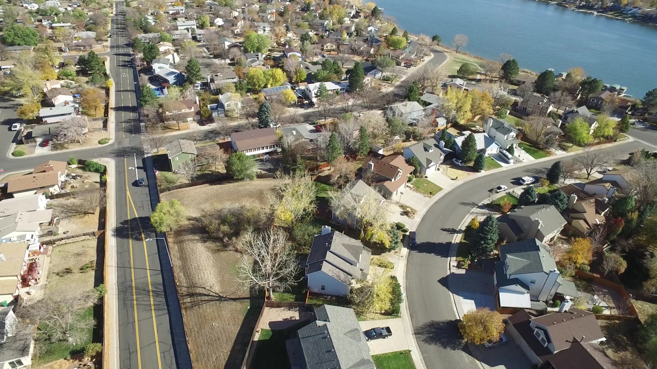 High aerial frame over Hidden Lake, Colorado showing a straight road on the left, a curved residential street, detached homes, fall trees, blue river water at the top, and no visible people.