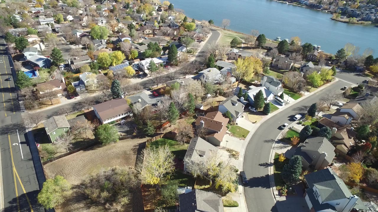 Second aerial frame over Hidden Lake, Colorado with more rooftops centered, a straight road along the left edge, yellow and bare trees, and no visible people.
