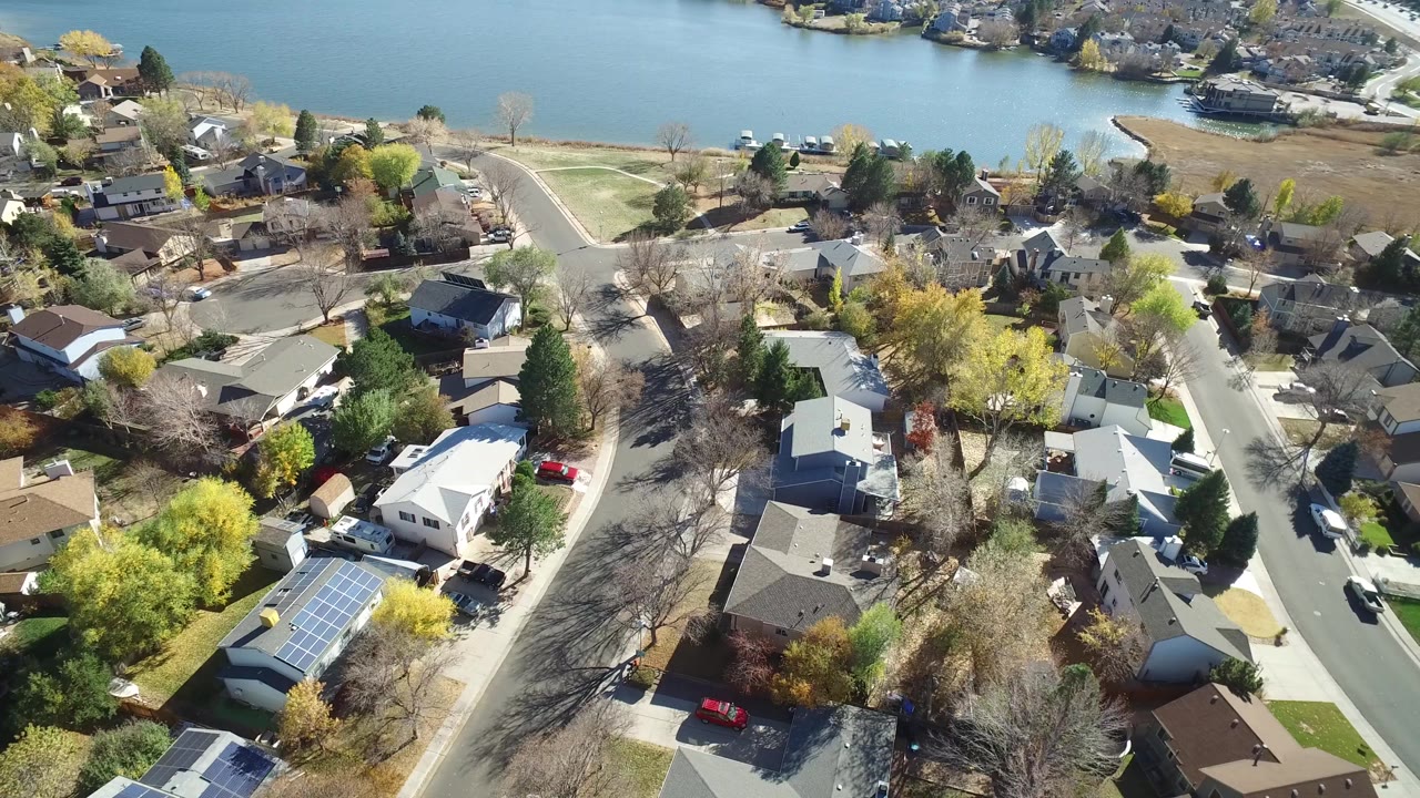 Aerial view in Hidden Lake, Colorado showing clustered rooftops, a pale curved road, tree shadows, and a lake shoreline running behind the neighborhood.