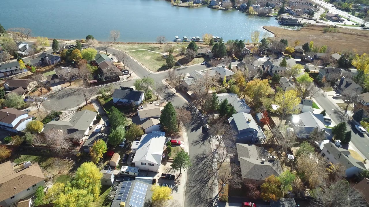 Second aerial frame over Hidden Lake, Colorado keeps the lake high in frame while curved residential streets, rooftops, yards, and yellow trees fill the foreground, with no visible people.