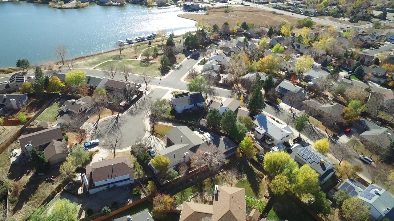 Aerial view in Hidden Lake, Colorado with sunlit lake water, a shoreline path, detached houses, fenced yards, and autumn-colored trees across the residential blocks.