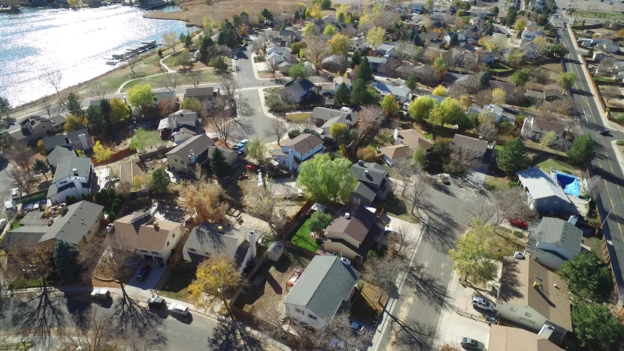 High aerial view over Hidden Lake, Colorado homes and curved streets, with a bright reflective lake along the upper left and no visible people.
