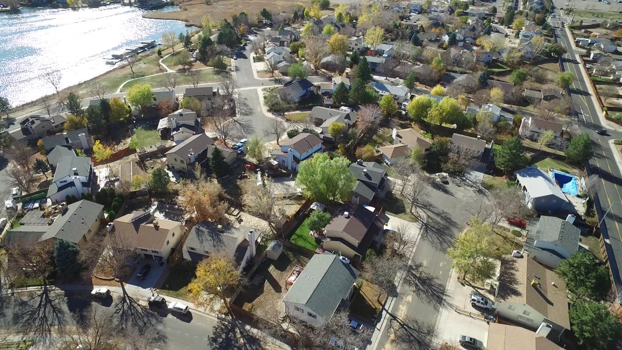 Aerial frame over Hidden Lake, Colorado showing rooftops, driveways, parked cars, leafless trees, green trees, and lake glare at the top left with no visible people.