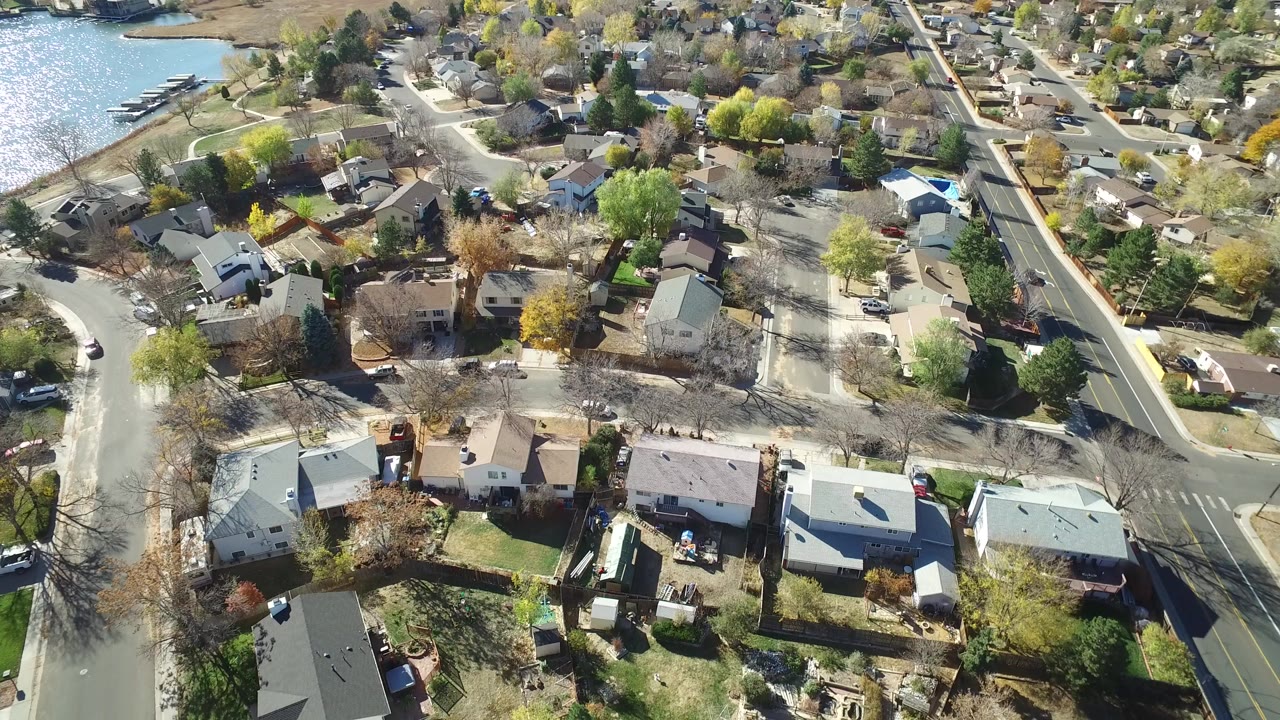Aerial frame over Hidden Lake, Colorado showing detached homes, intersecting residential roads, early-season trees, and water along the upper left edge, with no visible people.