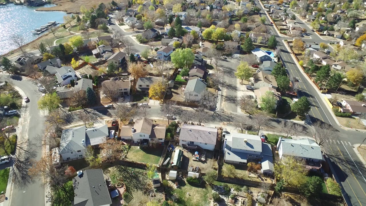 Aerial view in Hidden Lake, Colorado with light-colored rooftops, a curving street, roadside trees, and a strip of water near the top left, with no visible people.