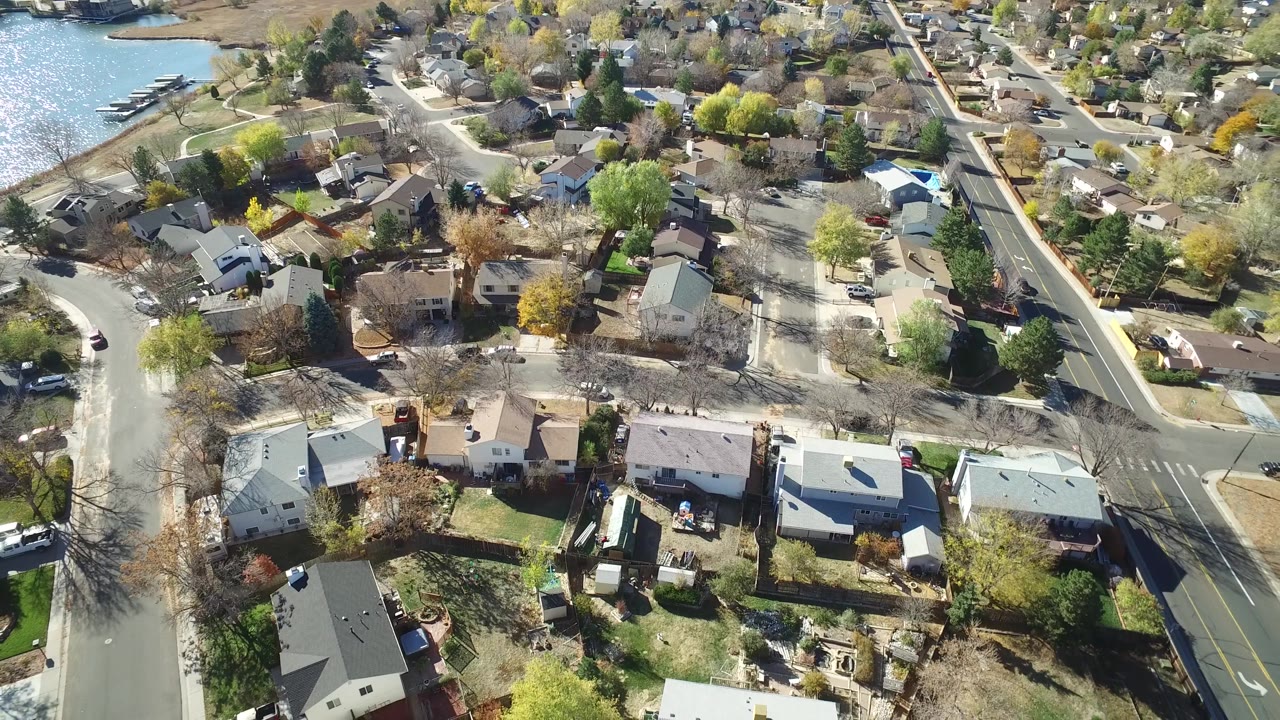 Oblique drone view of houses, an intersection, trees, and a road running along the right side.